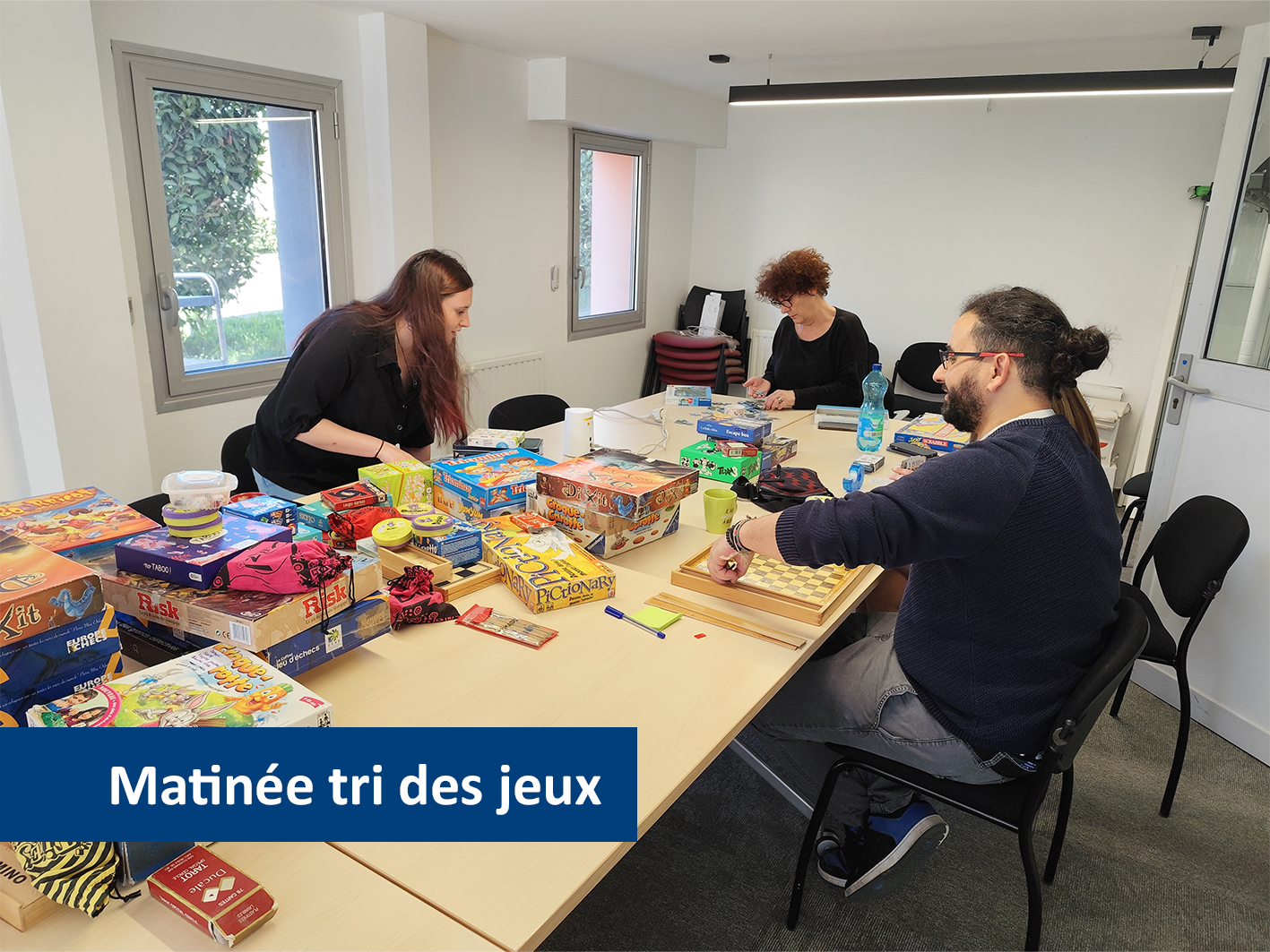 Trois personnes trient et vérifient des jeux de société dans une salle lumineuse, avec de nombreuses boîtes de jeux réparties sur deux grandes tables.